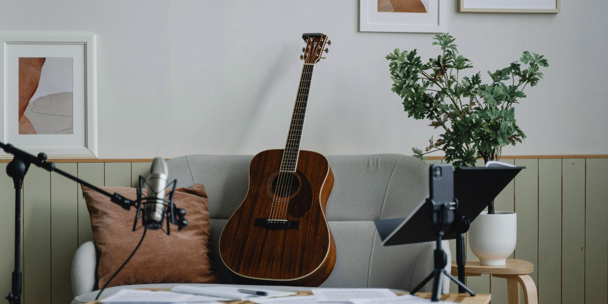 A guitar on a chair in a cosy music space featuring a microphone, music stand, plant, and framed artwork on the wall.