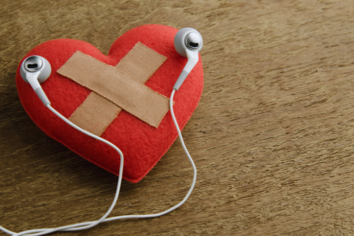A red fabric heart with two band-aids crossed on it sits on a wooden surface. Earbuds are placed on the heart.