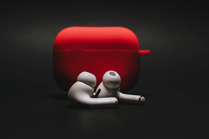 White wireless earbuds are placed in front of a red earbud case, on a dark background.