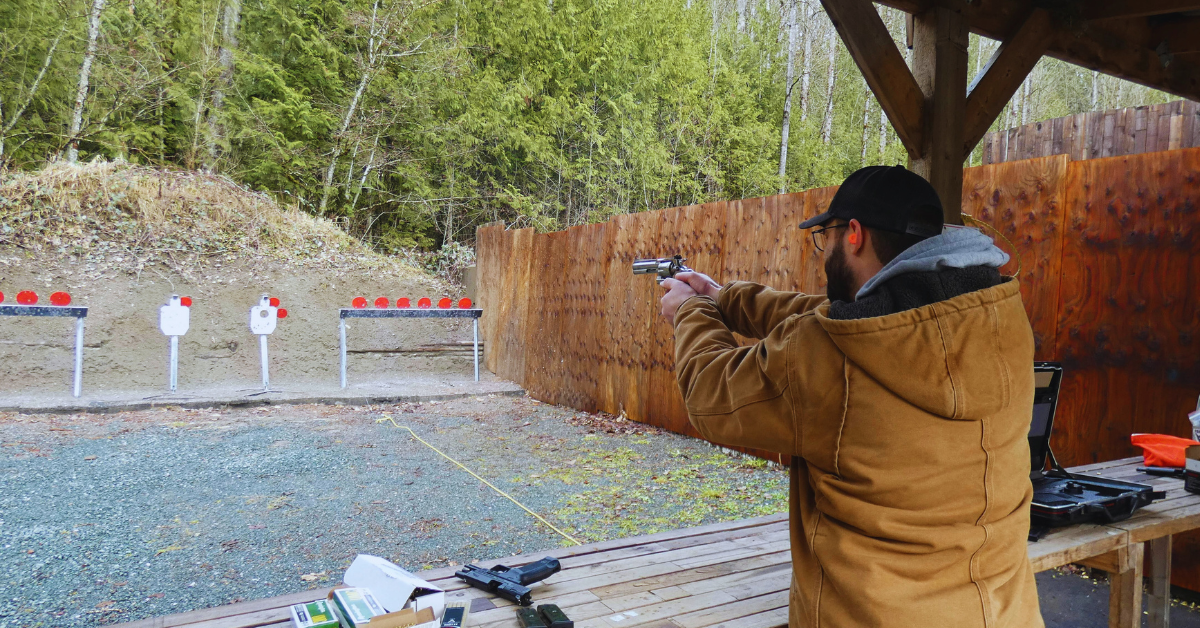 A man is on an outdoor shooting A man in a brown jacket and cap aims a pistol at red and white targets at an outdoor shooting range.