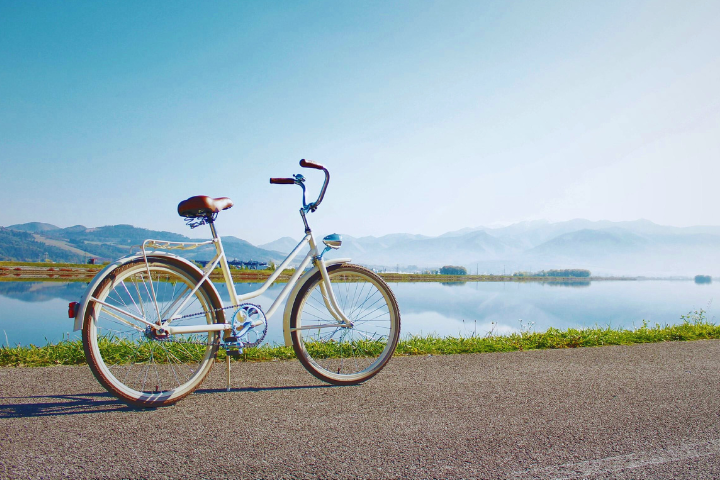 A white bicycle with a brown seat is parked on a path beside a serene lake, with mountains and a clear blue sky in the background, conveying tranquillity.