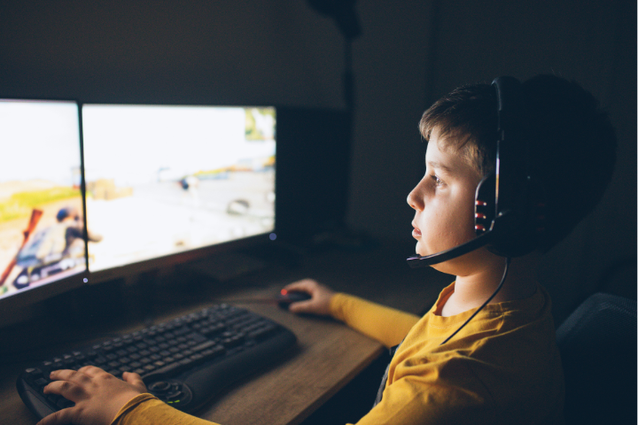 A boy wearing a headset intensely focuses on a video game displayed on dual monitors.