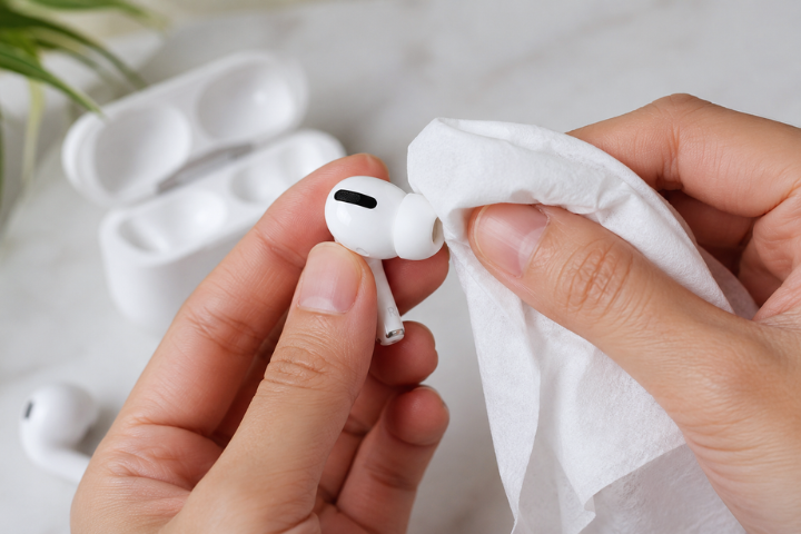 Hands cleaning wireless earbuds with a white cloth. An open charging case sits in the background on a marble surface.